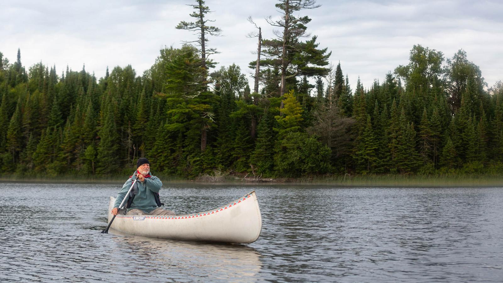 Man paddling a canoe in a lake surrounded by evergreen forest